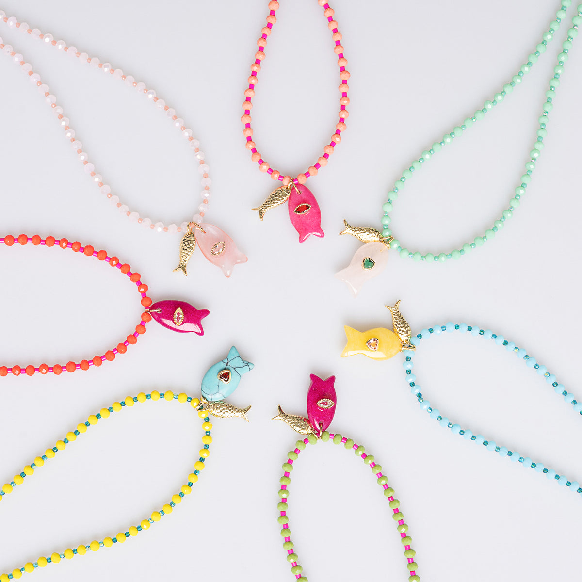 A set of colorful beaded necklaces arranged in a circle on a light gray background. Each necklace features a small fish-shaped charm with a gold tail and a tiny gemstone, paired with a string of beads in pink, mint green, coral, light blue, yellow, and purple.