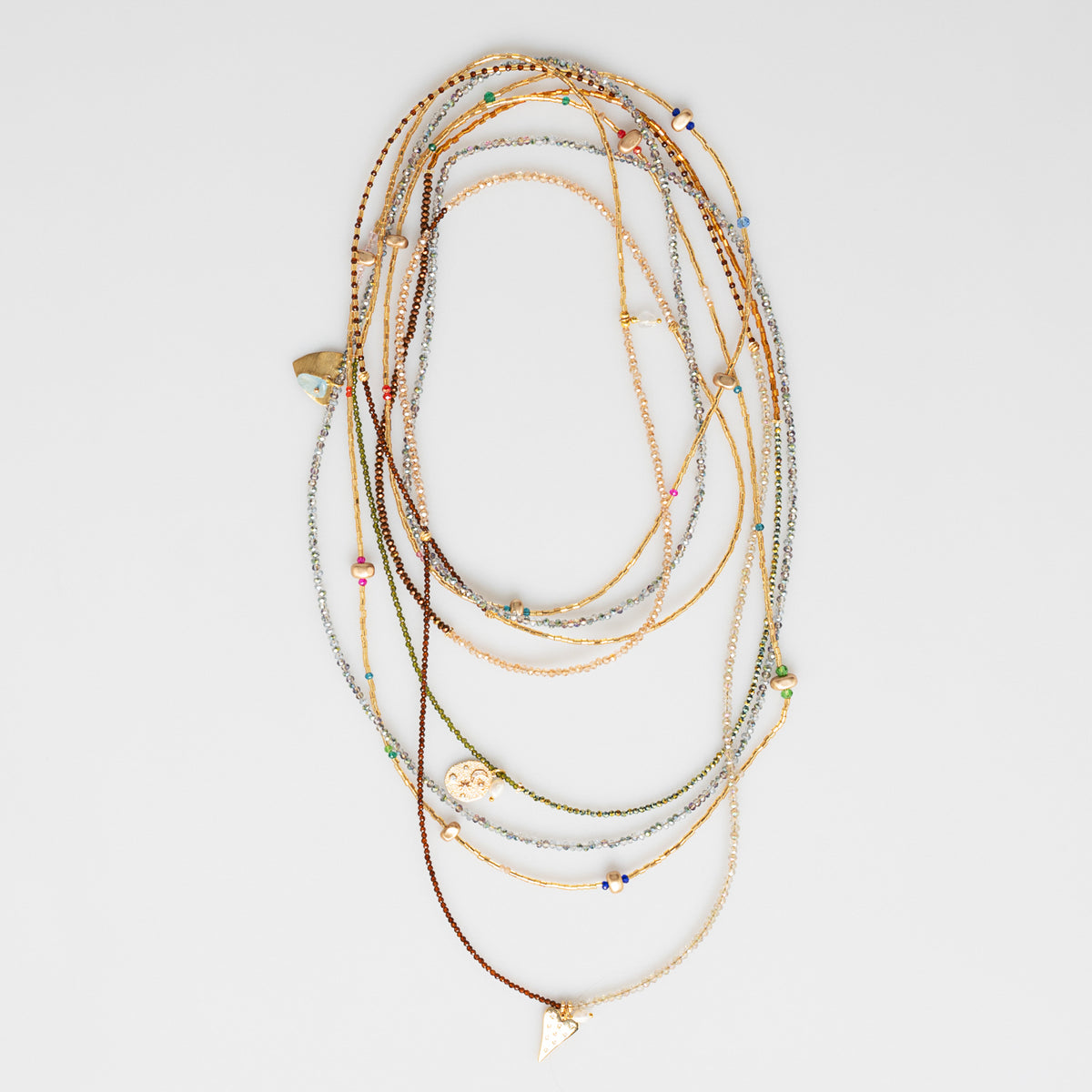 A stack of multi-strand bohemian-style beaded necklaces with gold, silver, and brown chains, small colorful beads, and charms, including heart charm, displayed on a light gray background.