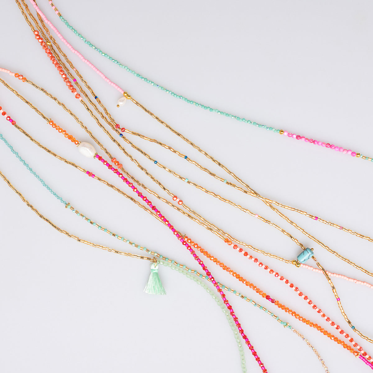 A set of bohemian-style beaded necklaces with gold-toned twisted cords, multicolor seed beads, and a small mint green tassel charm, arranged diagonally on a light gray background.